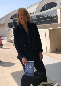 Anastasia standing at the curb of the airport holding a book that says CONGO on the front.
