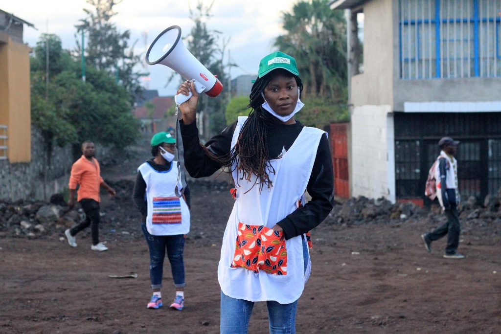 Teen Congolese mom holding up bull horn and wearing PPE the girls have put together themselves.