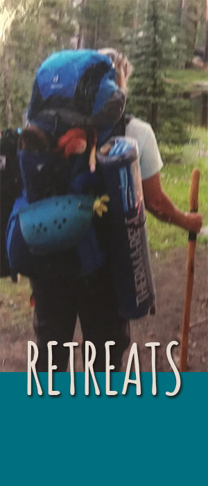 Anastasia from behind wearing a backpck and a walking stick in hand. She is looking out from trail to a lake.